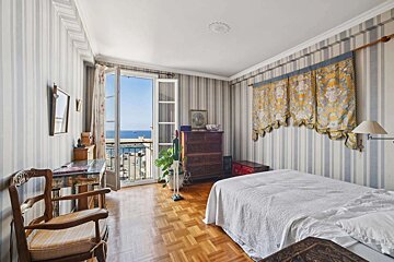 A traditional bedroom with striped wallpaper and parquet floors, featuring a balcony door open to a picturesque sea view. Furnished with a bed, dresser, and desk.
