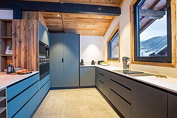 Modern chalet kitchen with blue-gray cabinets, a wooden ceiling, and windows showing a snowy mountain landscape.