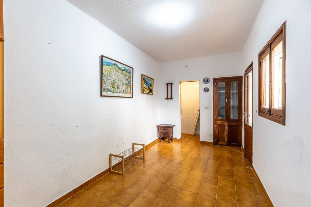 A bright room with white walls, brown tiled floor, and dark wood furniture, including a cabinet, small table, and shoe rack. Paintings adorn the walls.