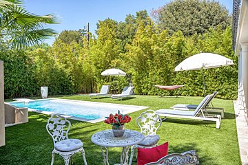 A sunny backyard features a blue swimming pool, green lawn, lounge chairs, a white patio set, and a hammock, surrounded by lush bamboo and trees.