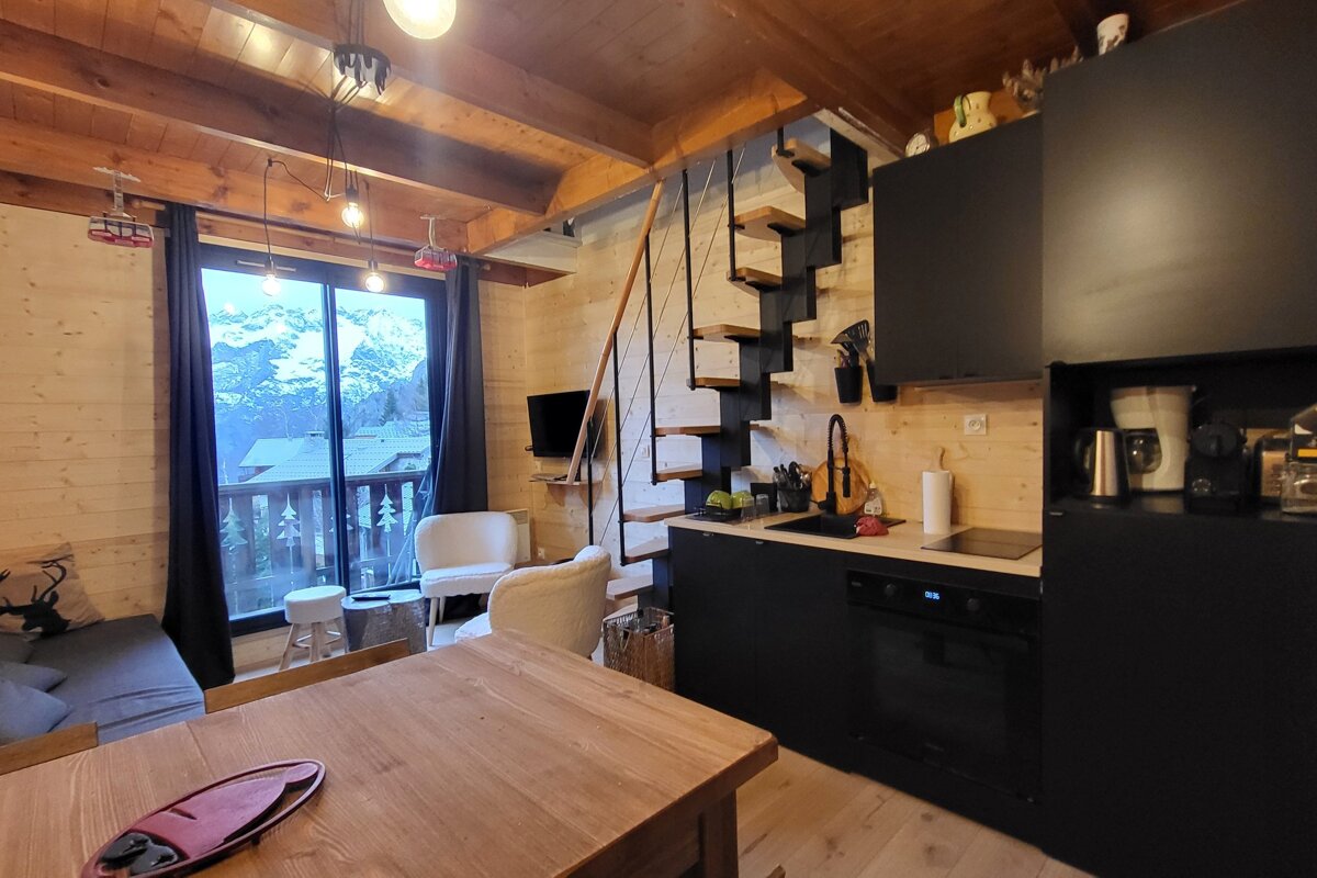 A cozy wooden chalet interior features a modern black kitchen, a spiral staircase, and large windows with a stunning mountain view.