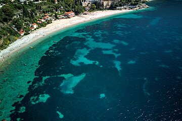 Aerial shot of a serene coastline with a sandy beach, hillside homes, and stunning clear turquoise merging into deep blue ocean water.