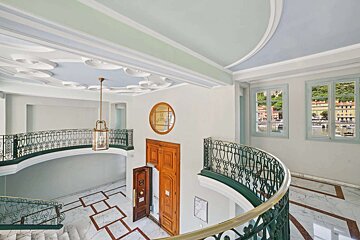 Grand interior with ornate green and gold staircase, marble floors, decorative ceilings, and windows overlooking a colorful coastal town.