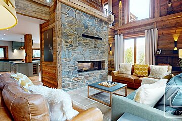 Rustic, open-plan living room with a large stone fireplace, wooden chalet-style decor, cozy sofas, and a modern kitchen visible beyond.