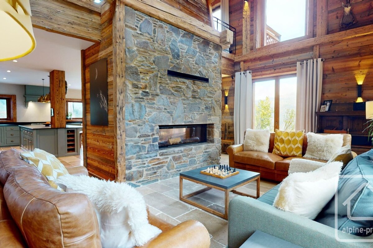 A rustic chalet living room with a stone fireplace, cozy sofas, a chess table, wooden beams, and a view of the kitchen in the background.