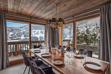 A rustic dining room with a wooden table, antler chandelier, and large windows overlooking a snowy mountain landscape.