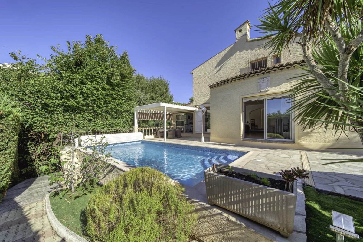 A beige villa features a private swimming pool with stone tiling, surrounded by lush green trees and plants under a blue sky.