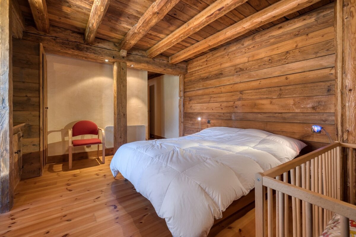 Cozy chalet bedroom with rich wooden walls, floor, and ceiling beams. Features a large bed with white duvet, red chair, and a baby crib.