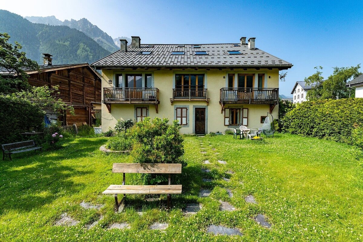 A charming yellow house with balconies and a green lawn featuring a wooden bench, set against majestic mountains under a bright blue sky.
