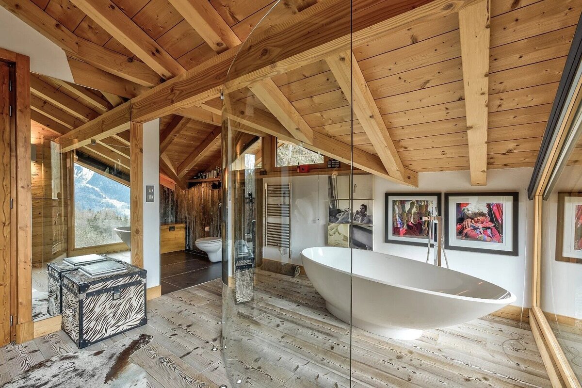 Wood-clad attic bathroom with a white freestanding tub, glass shower, animal print trunk, and panoramic mountain window.