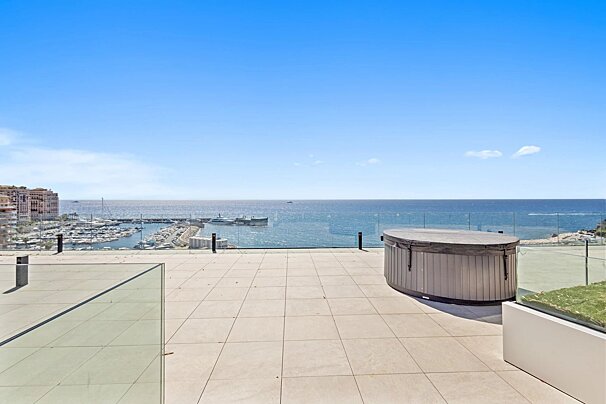 Tiled rooftop terrace with a hot tub and glass railings, offering a panoramic ocean and harbor view under a bright blue sky.