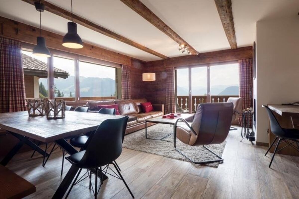 Cozy alpine cabin interior with a wooden dining table, leather seating, exposed ceiling beams, and scenic mountain views.