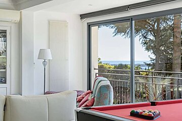 A bright room with a red pool table and a panoramic view of the sea and trees through large glass doors, offering an inviting space.