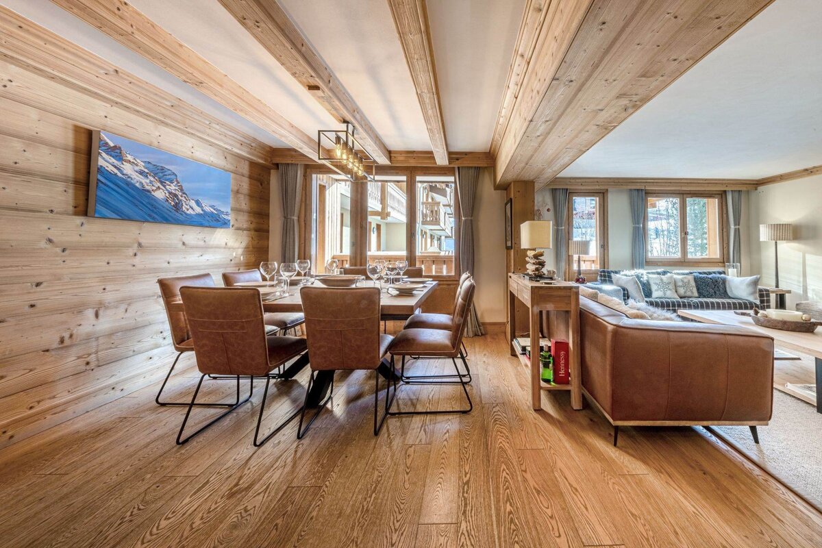 A warm, open-plan chalet-style living space with extensive wooden paneling, a kitchen bar with woven stools, and a dining area.