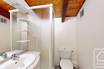 A compact bathroom with a marble-top sink, mirrored cabinet, frosted glass shower, toilet, and rustic wooden beamed ceiling.