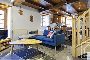 A cozy, open-plan living room and kitchen featuring a blue L-shaped sofa, wooden beamed ceiling, and a wooden staircase.