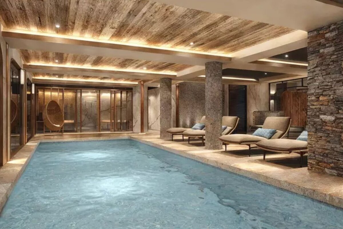 Luxurious indoor pool area with stone pillars, a wood-beamed ceiling, warm ambient lighting, and poolside lounge chairs.