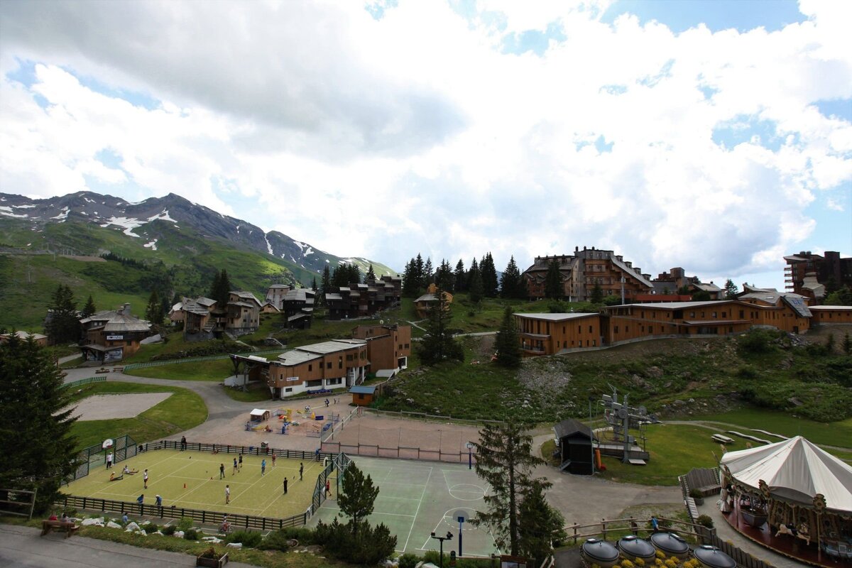 Alpine village resort with people playing on sports courts, diverse buildings nestled on green hills, and snow-capped mountains under a cloudy sky.
