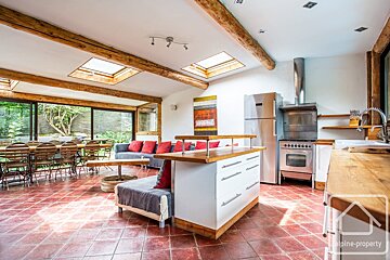 Spacious, bright open-plan kitchen, dining, and living room with red tiled floors, wooden beams, skylights, and large windows to a garden.