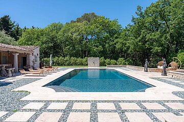 A spacious outdoor pool with a traditional house, lounge chairs, and decorative statues on stone paving, surrounded by lush green trees and a clear blue sky.