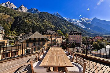 A rooftop terrace with a wooden dining set provides panoramic views of a charming town and majestic snow-capped mountains under a clear blue sky.