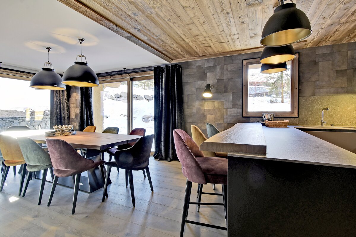 A chic chalet dining room & kitchen island featuring colorful velvet chairs, a wooden ceiling, and large windows with a snowy view.