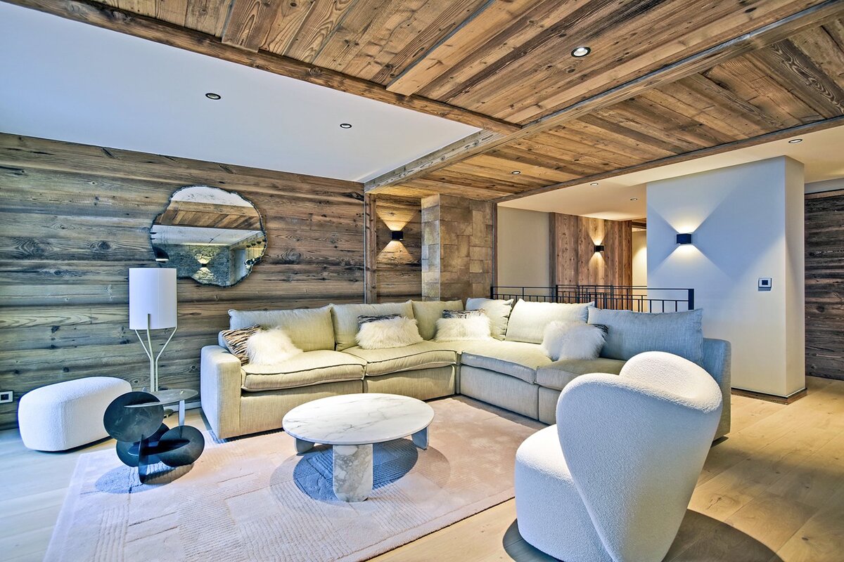 Luxurious chalet living room with rich wood walls & ceiling, large L-shaped sofa, modern marble coffee table, and stylish white seating. Warm ambiance.