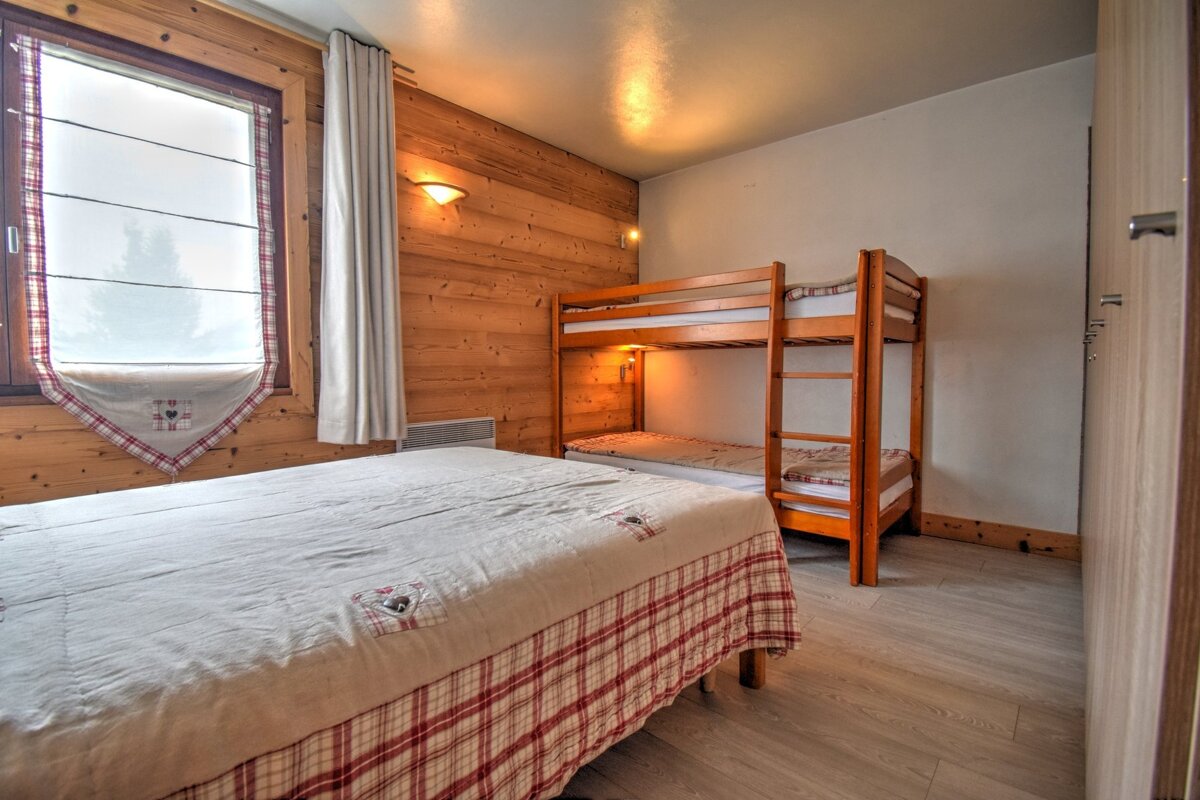 A cozy, rustic bedroom with wood-paneled walls, featuring a large bed and wooden bunk beds. A window with a decorative blind and warm wall lights.