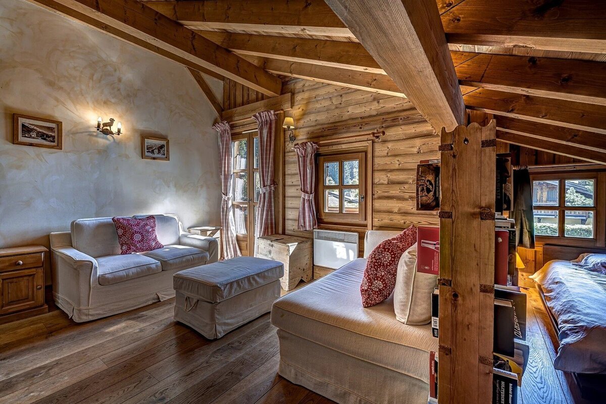 A cozy, rustic attic room with wood-paneled walls, exposed beams, and a blend of living and sleeping spaces. Warm lighting and windows complete the scene.
