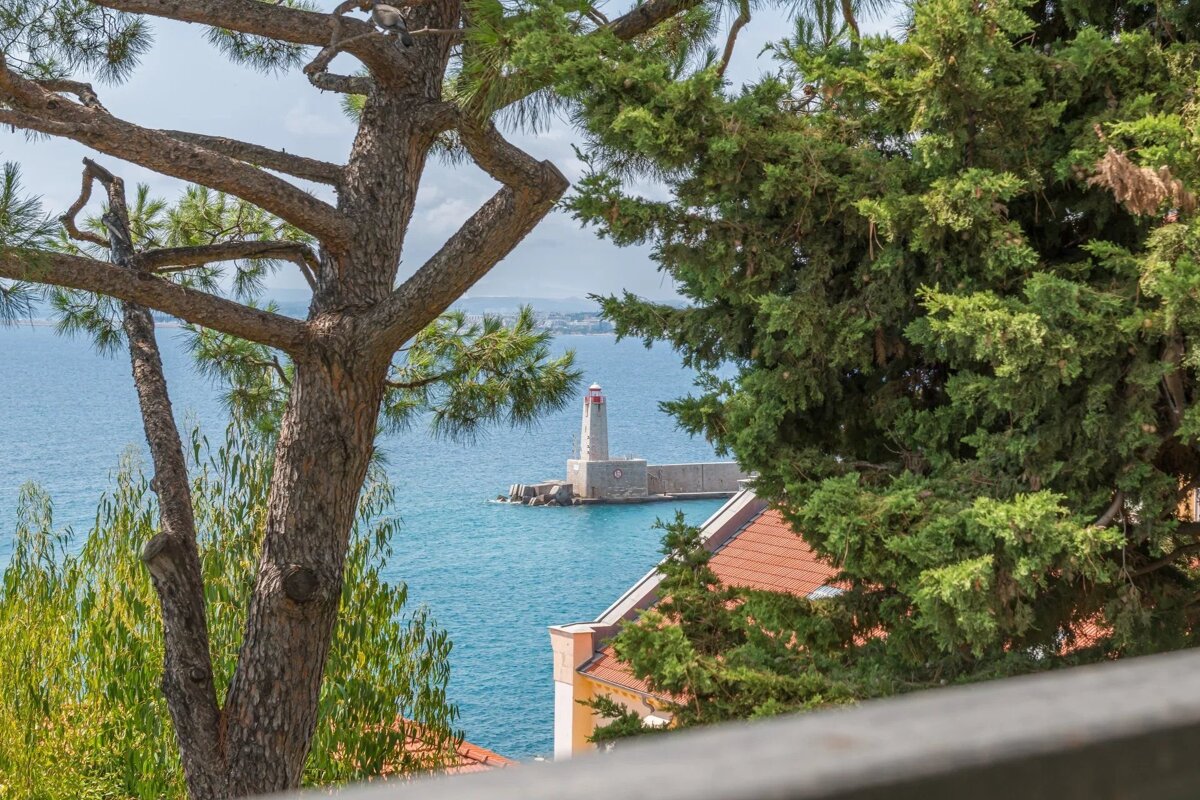 A scenic view of a white lighthouse on a pier in blue water, framed by green trees and with rooftops below.
