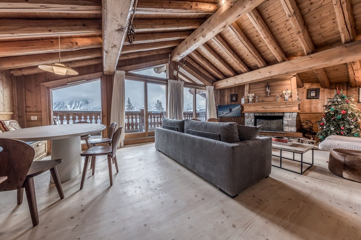 Spacious wooden chalet living room with a festive Christmas tree, stone fireplace, and a stunning snowy mountain vista.