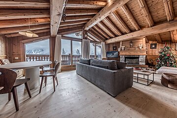 Spacious wooden chalet living room with a festive Christmas tree, stone fireplace, and a stunning snowy mountain vista.