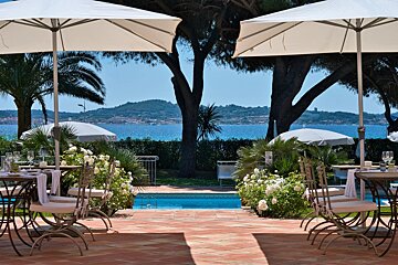 Outdoor dining area with white umbrellas, set tables, and a small pool, overlooking a stunning blue sea and distant green hills.