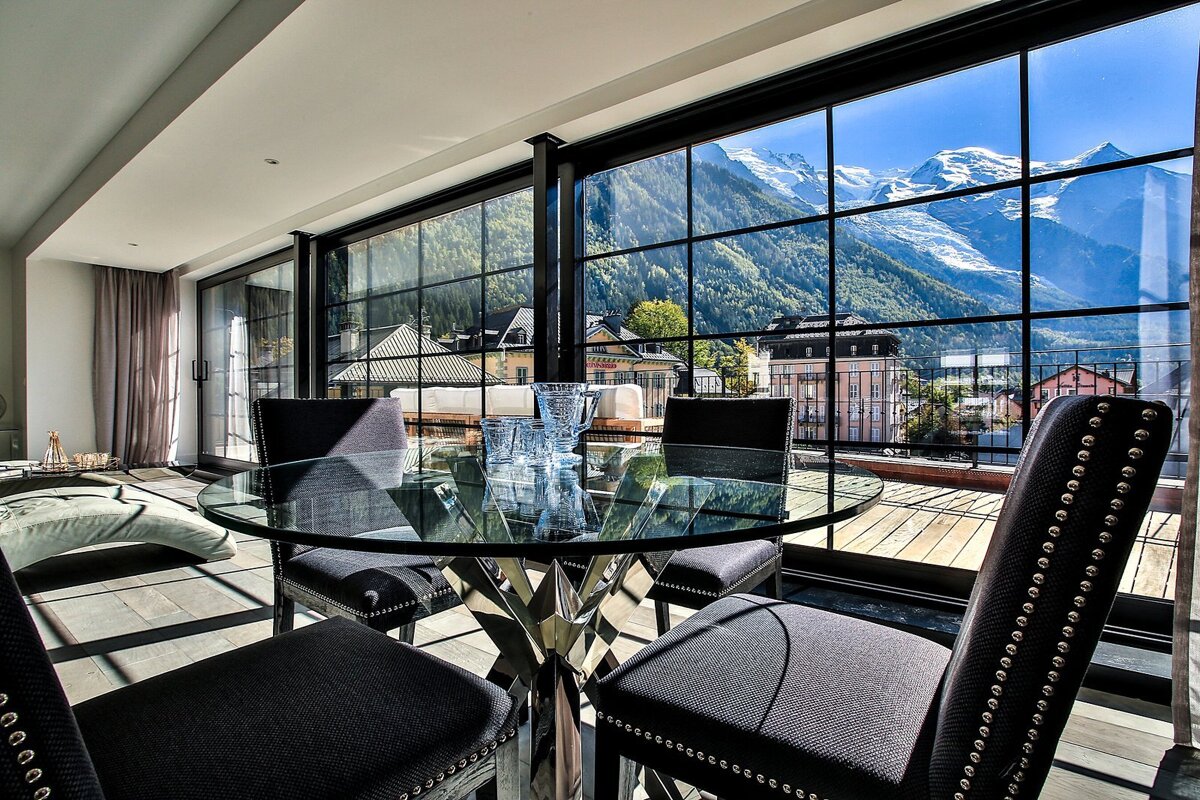 Chic interior with a glass table and black chairs, boasting panoramic windows showcasing snow-capped mountains and a town.