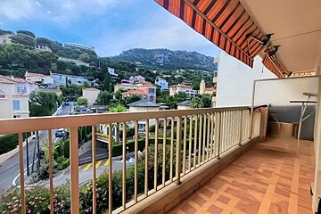 A balcony view over a charming hillside town with houses, a winding road, and green trees, all set against a backdrop of distant mountains.