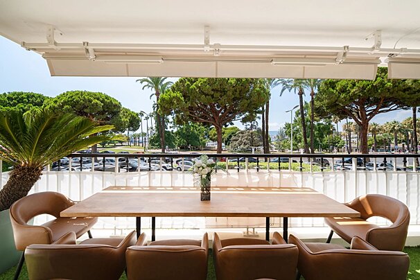 Sunny balcony terrace featuring a wooden dining table with brown chairs, overlooking palm trees and lush greenery.