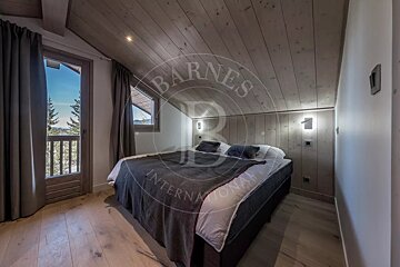 A charming, modern bedroom featuring a wooden sloped ceiling, light wood floor, and a bed with dark bedding. A door opens to a balcony with a scenic mountain view.