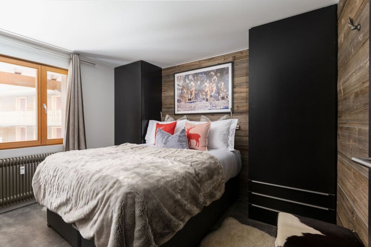 A cozy chalet-style bedroom featuring a plush bed flanked by tall black wardrobes against a rustic wood-paneled accent wall.