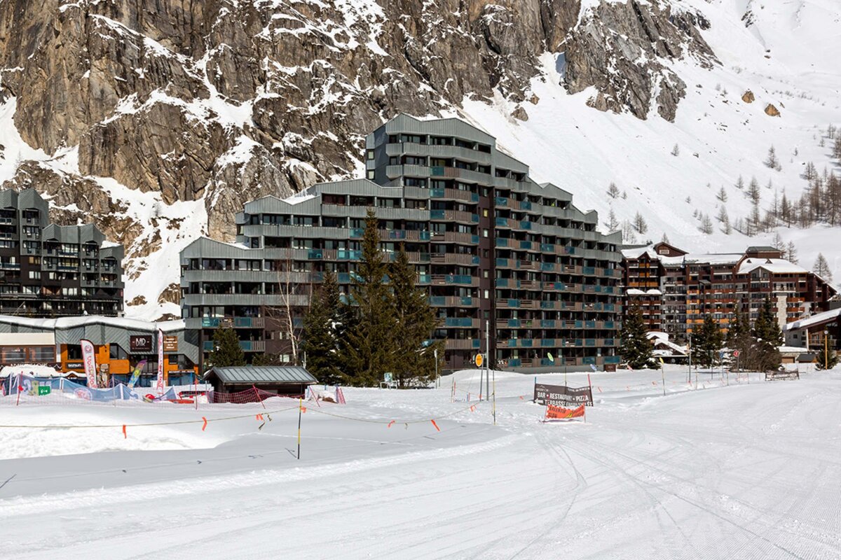 A snowy ski resort scene featuring large apartment buildings, pine trees, and a ski slope, all framed by rugged, snow-dusted mountains.