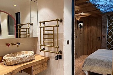 A rustic bathroom features a unique stone sink, wooden counter, brass fixtures, and a towel warmer, with a glimpse into a cozy, wood-paneled bedroom.
