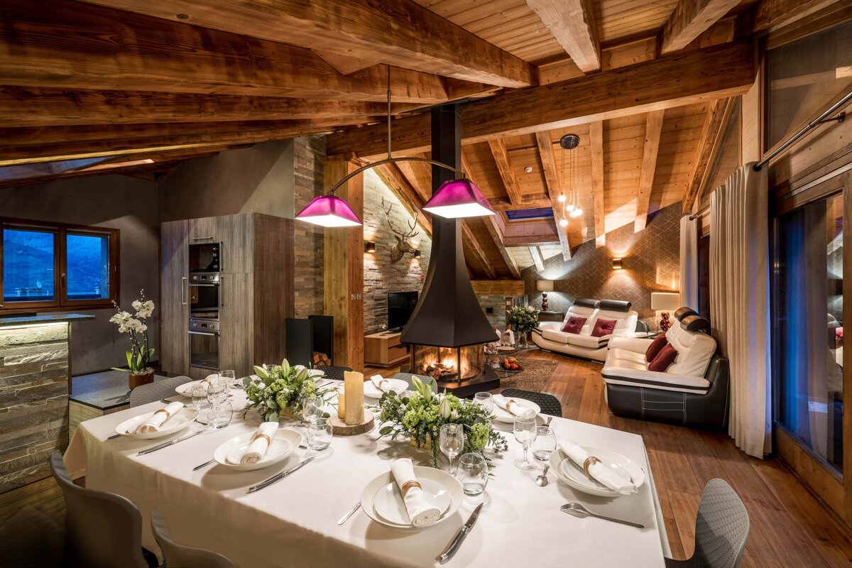 Luxurious chalet interior featuring a set dining table, rustic wooden beams, a central fireplace, and a cozy living area.