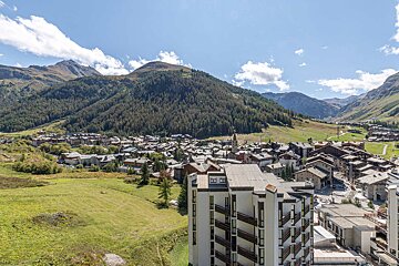 High-angle view of an alpine town in a green valley surrounded by forested mountains, with a modern building in the foreground.