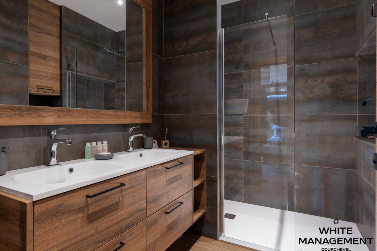 A modern bathroom featuring a double wood vanity with white sinks, dark tiled walls, and a clear glass shower enclosure.