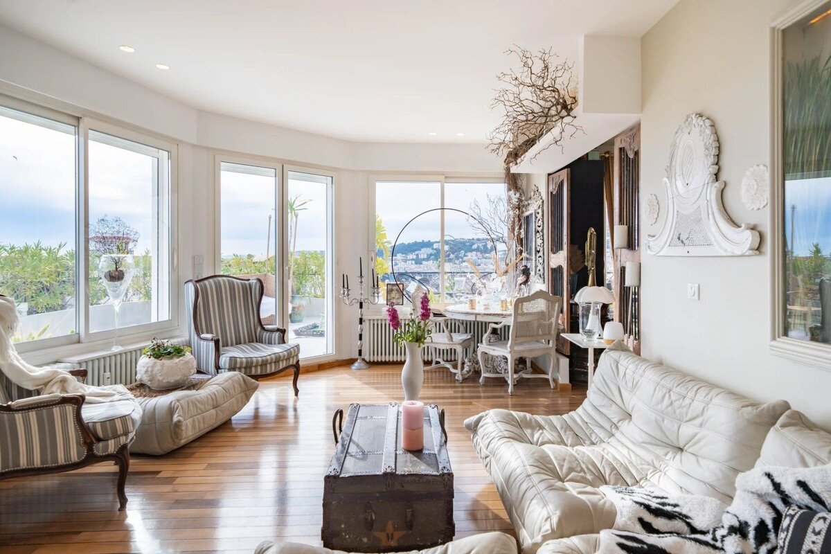 A bright, eclectic living room with large windows, city views, hardwood floors, white sofas, striped chairs, and ornate, vintage-style decor.