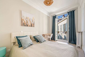 A bright, modern bedroom features a large bed with blue cushions, abstract art, a desk, and an open balcony overlooking charming buildings.