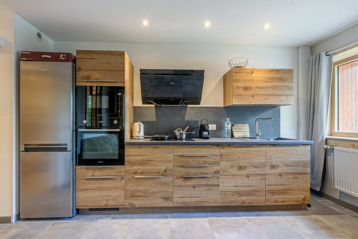 A modern kitchen with light wood cabinets, stainless steel fridge, built-in oven, dark counters, and a window on the right.