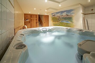An indoor hot tub with still water and soft lighting, set against a mountain mural, wooden doors, and tiled walls.