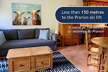 Cozy alpine interior with a grey sofa, wooden coffee table, cow art, and promotional text about being near the Prarion ski lift.