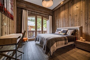 Rustic chalet bedroom with distressed wooden walls, a cozy bed, desk, and a balcony view of trees and other chalets.