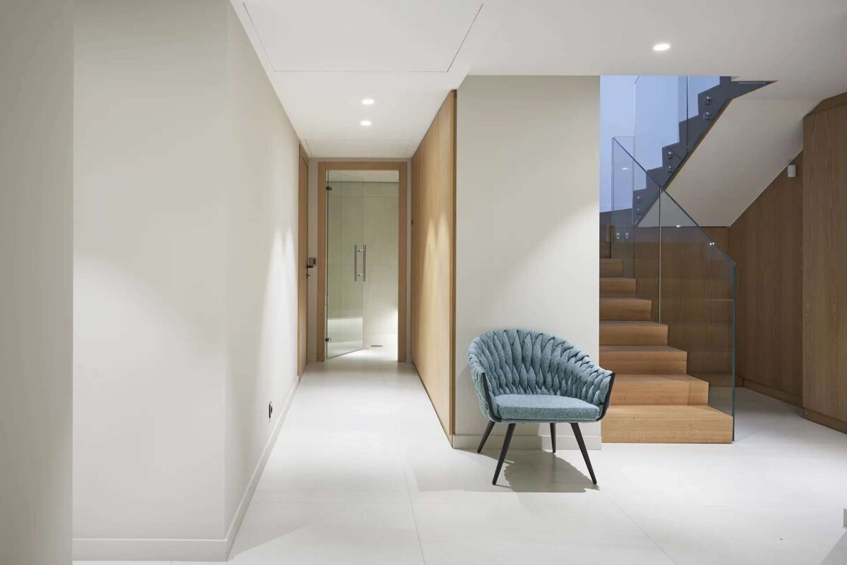 A clean, modern interior features a light blue armchair by wooden stairs with a glass railing. A white corridor leads to a distant glass door.